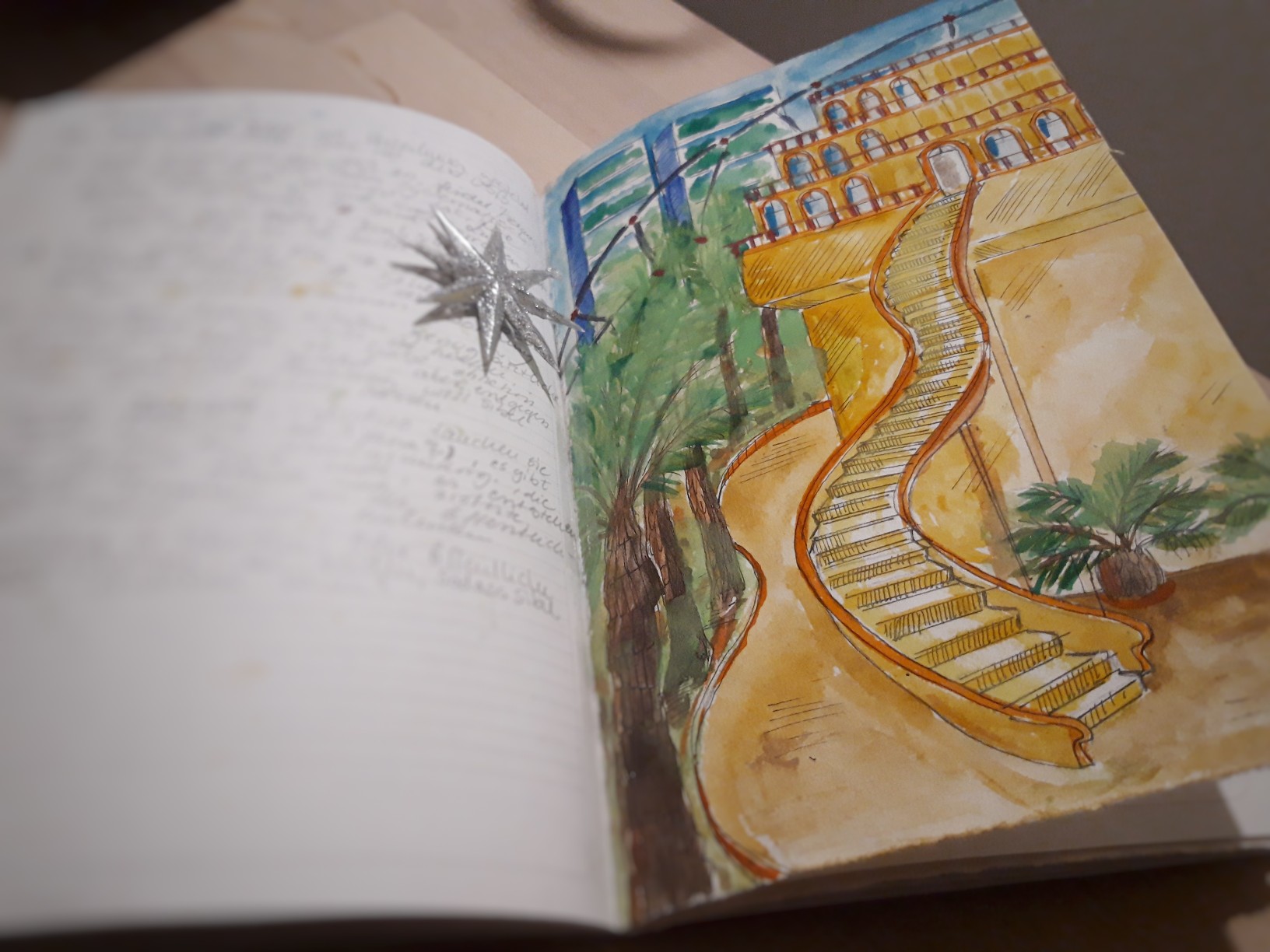 Aufgeschlagene Seite im Notizbuch mit einer farbigen Aquarellzeichnung mit einem gelben Gebäude in einem Palmengarten
