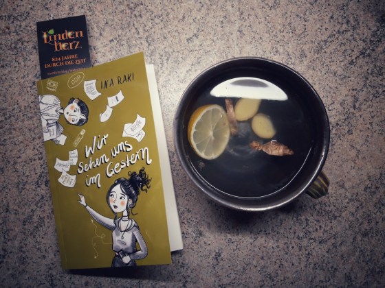 Buch "Wir sehen uns im Gestern" von Ina Raki neben einer Tasse Tee mit Ingwer und Zitrone