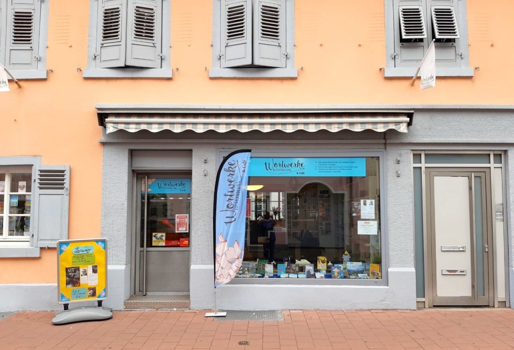 Die Wortwerke-Buchhandlung in Rastatt - davor eine blaue Fahne, ein Aufsteller und im Schaufenster sieht man Bücher.