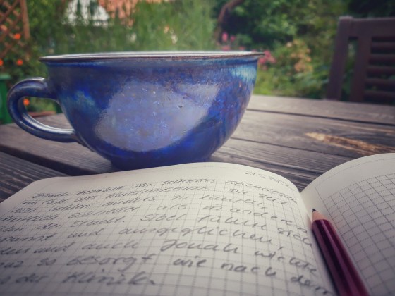 aufgeschlagenes Notizbuch im Vordergrund, dahinter eine blaue Keramiktasse und Ausblick auf den Garten