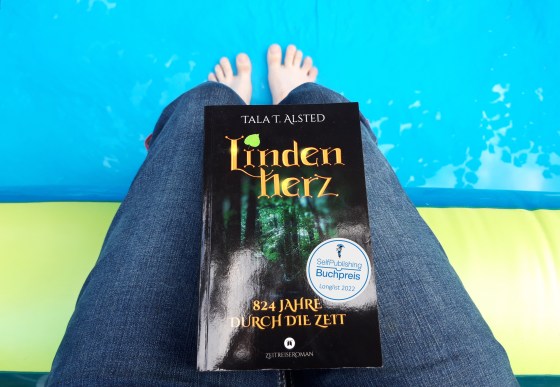 Buch "Lindenherz - 824 Jahre durch die Zeit" von Tala T. Alsted auf dem Schoß, dazu der Longlist-Batch des SelfPublishing Buchpreis