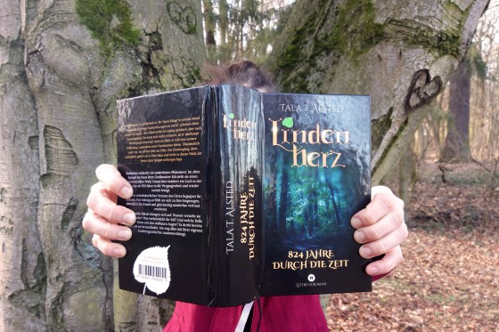 Tala T. Alsted hält ihr Buch "Lindenherz - 824 Jahre durch die Zeit" in die Kamera - im Hintergrund ein Baum