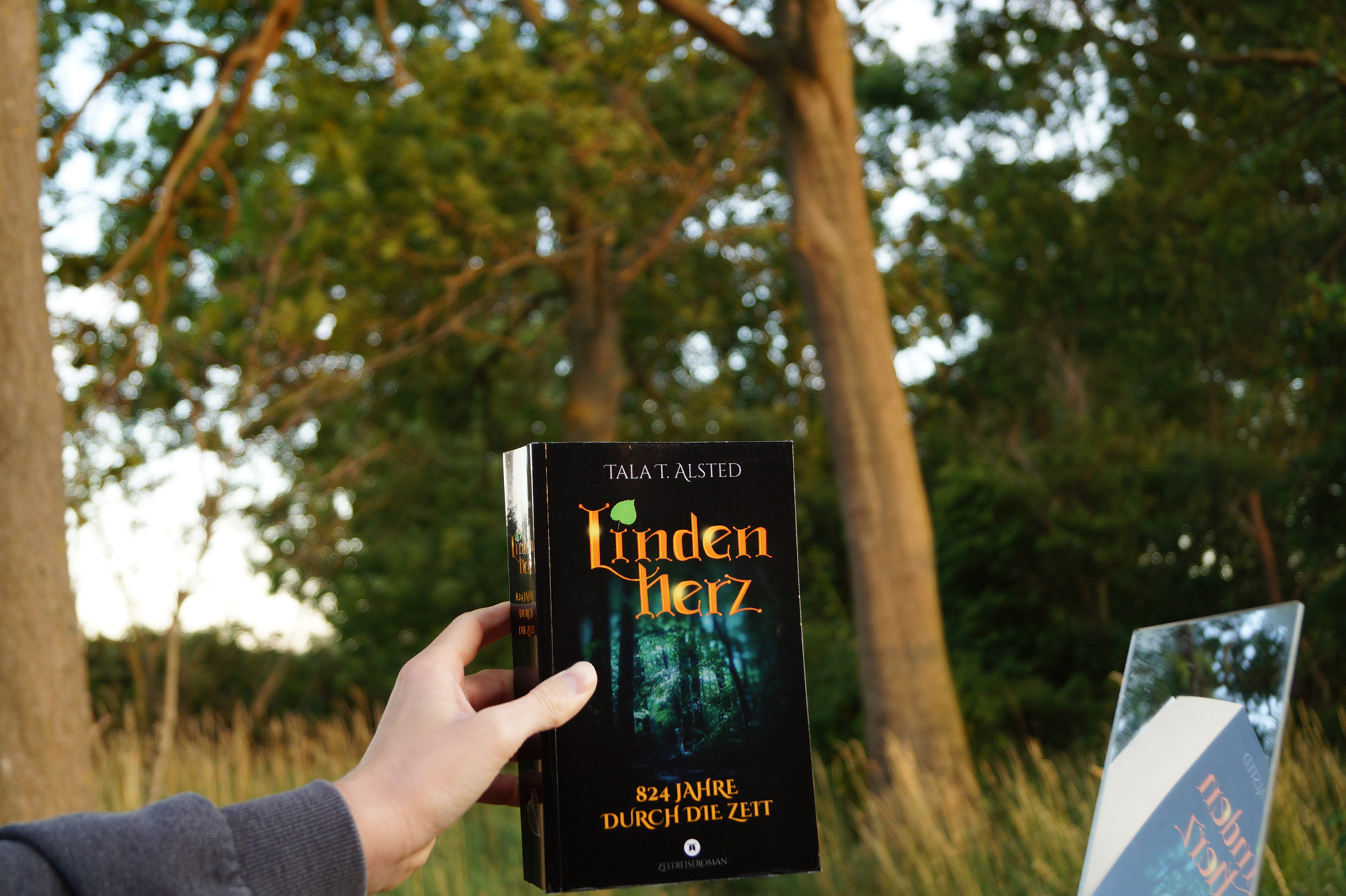 Jemand hält das Buch "Lindenherz" von Tala T. Alsted an einen Spiegel. Im Hintergrund ist Wald zu sehen.