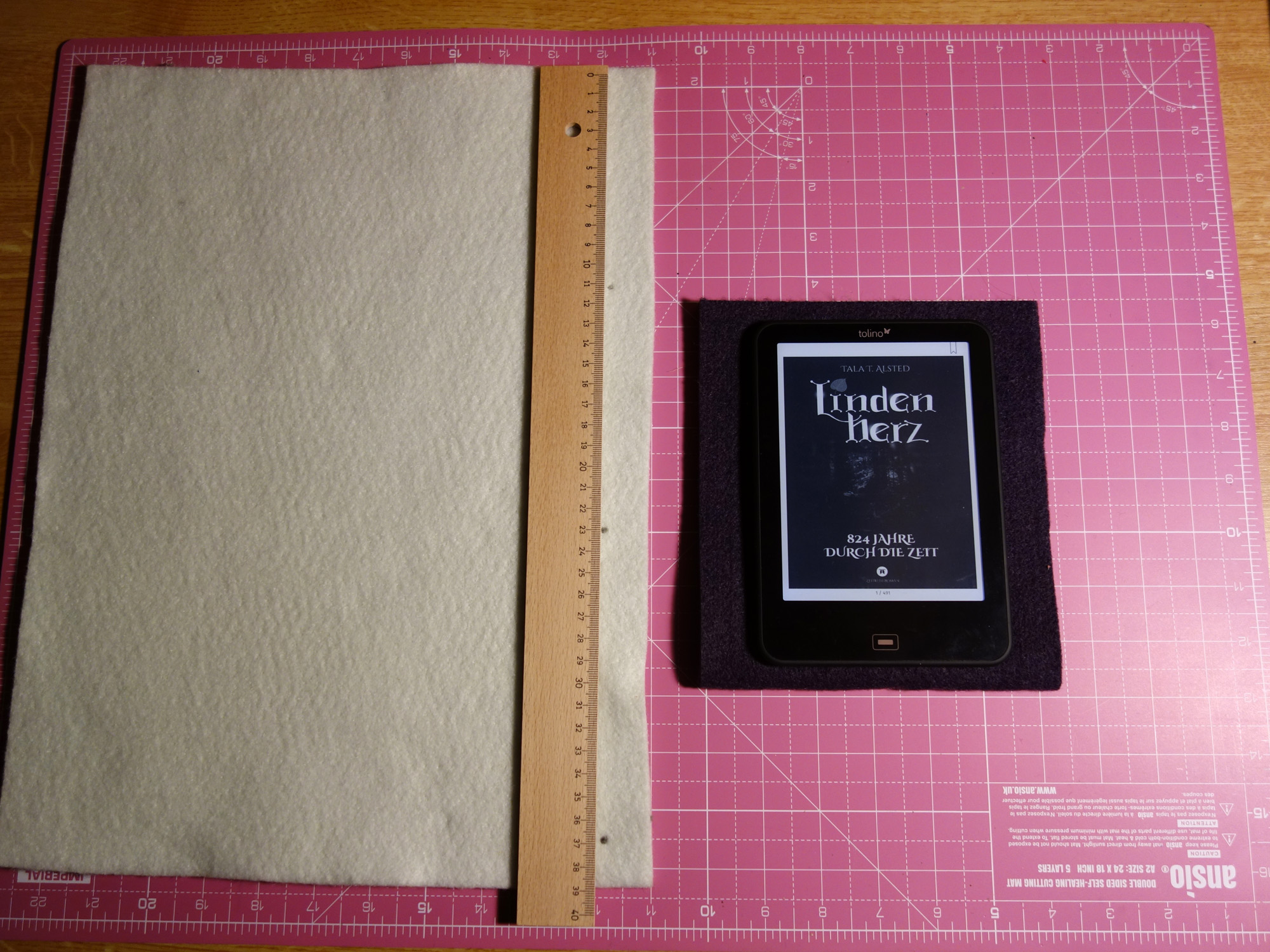 Schneidematte mit weißem und violettem Filz, einem Lineal und dem E-Book-Reader, für den die Hülle genäht werden soll. Auf dem Reader ist das Cover von "Lindenherz - 824 Jahre durch die Zeit" von Tala T. Alsted zu sehen.