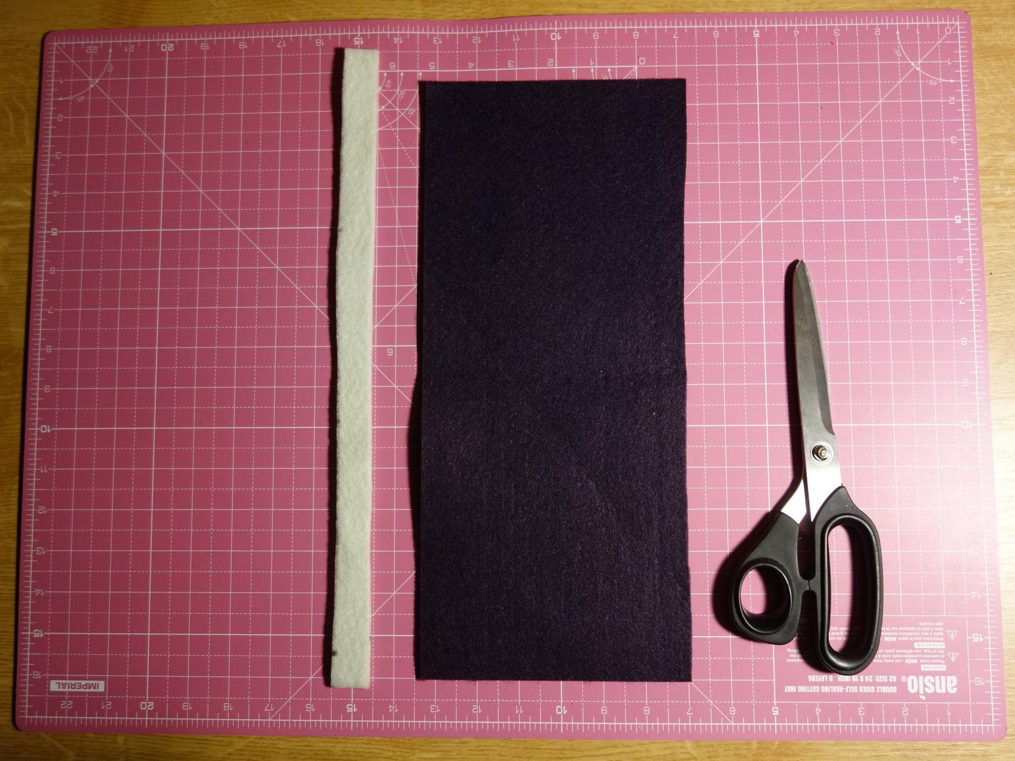 Ein langer, dünner weißer Stoffstreifen liegt neben einem rechteckigen violetten Stoffstück und einer Schere.