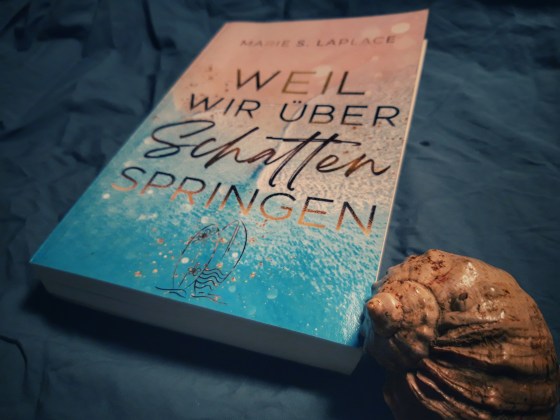 Das Buch "Weil wir über Schatten springen" von Marie S. Laplace liegt auf blauem Grund und davor eine Muschel