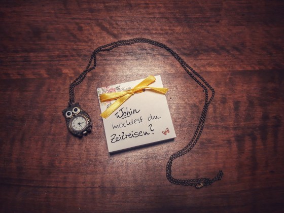Ein Notizblock mit Schleife, auf dem steht "Wohin möchtest du Zeitreisen?" liegt auf einem Tisch, darum herum eine Uhr in Eulenform an einer Kette