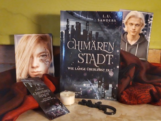 Buch "Chimärenstadt" von L.U. Sanders und zwei Charakterpostkarten, ein Lesezeichen, ein Teelicht und eine Echse aus Holz